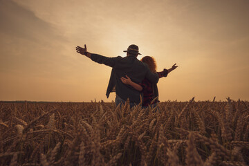 Happy man and woman are standing in their agricultural field in sunset. They are cultivating wheat...