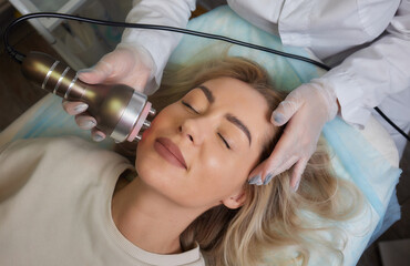 A cosmetologist in rubber gloves does the rf-lifting procedure on the face. In the background, the...