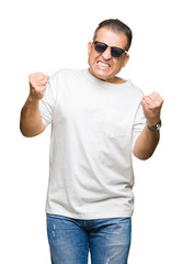 Naklejka premium Middle age arab man wearig white t-shirt and sunglasses over isolated background very happy and excited doing winner gesture with arms raised, smiling and screaming for success. Celebration concept.