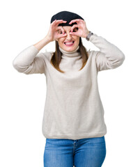 Middle age mature woman wearing winter sweater and beret over isolated background doing ok gesture like binoculars sticking tongue out, eyes looking through fingers. Crazy expression.