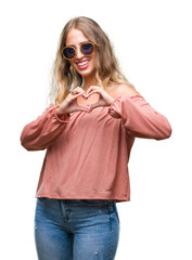 Beautiful young blonde woman wearing retro sunglasses over isolated background smiling in love showing heart symbol and shape with hands. Romantic concept.