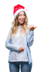 Beautiful young blonde woman wearing christmas hat over isolated background smiling with happy face looking and pointing to the side with thumb up.