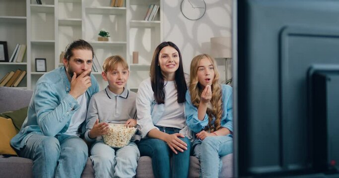 Young caucasian parents with their children are sitting on the sofa in the living room, eating popcorn and watching TV.