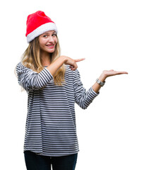 Young beautiful blonde woman wearing christmas hat over isolated background amazed and smiling to the camera while presenting with hand and pointing with finger.