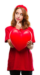 Young beautiful woman holding red heart in love over isolated background serious face thinking about question, very confused idea