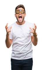 Young handsome man wearing carnival mask over isolated background crazy and mad shouting and yelling with aggressive expression and arms raised. Frustration concept.