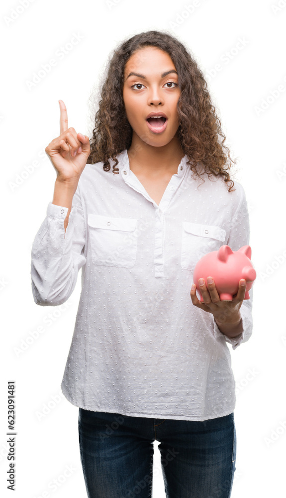 Sticker young hispanic woman holding piggy bank surprised with an idea or question pointing finger with happ