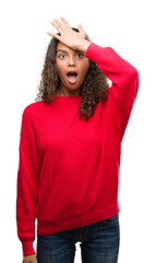 Young hispanic woman wearing red sweater surprised with hand on head for mistake, remember error. Forgot, bad memory concept.