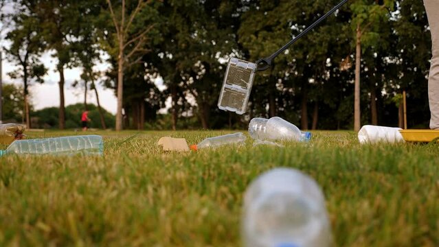 Close-up of a female activist collecting plastic garbage with tongs on the green grass. Volunteers help to save nature from plastic waste pollution.