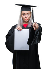Young blonde woman wearing graduate uniform holding degree over isolated background pointing with...