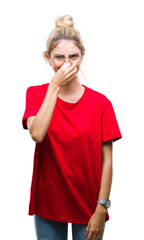 Young beautiful blonde woman wearing red t-shirt and glasses over isolated background smelling something stinky and disgusting, intolerable smell, holding breath with fingers on nose