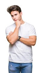 Young handsome man wearing white t-shirt over isolated background with hand on chin thinking about question, pensive expression. Smiling with thoughtful face. Doubt concept.