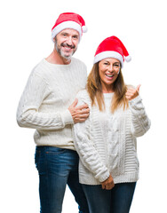 Middle age hispanic couple wearing christmas hat over isolated background smiling with happy face looking and pointing to the side with thumb up.