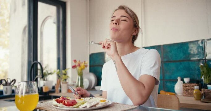 A blonde girl in a white t-shirt is enjoying her domestic breakfast in the kitchen on a sunny morning. Proper nutrition is the basis of a happy life
