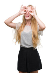 Obraz na płótnie Canvas Blonde teenager woman wearing moles shirt doing ok gesture like binoculars sticking tongue out, eyes looking through fingers. Crazy expression.