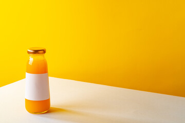 Glass bottle of orange juice on white table against yellow background
