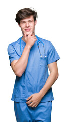 Young doctor wearing medical uniform over isolated background looking confident at the camera with smile with crossed arms and hand raised on chin. Thinking positive.