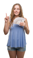 Obraz premium Young blonde woman holding dollars surprised with an idea or question pointing finger with happy face, number one