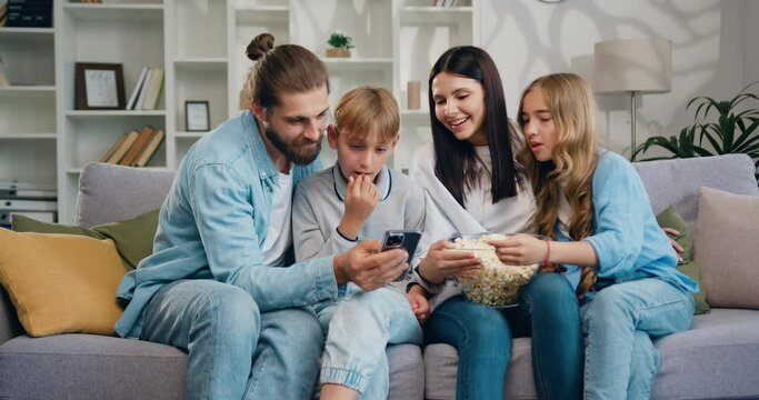 Parents father mother and cute little kids children watch funny social media video on mobile phone. Young family using modern wireless tech watching vlog, enjoy free time on internet.