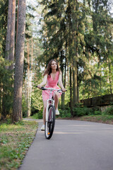 young active woman in pink dress rides a bicycle in park among the trees. Women fun weekend sports and recreation. Walking along bike path in national park, freedom and speed. riding