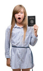 Young blonde toddler holding italian passport scared in shock with a surprise face, afraid and excited with fear expression