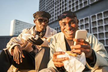 Cheerful multiracial hipster guys eating fast food and using smartphone while sitting on street stairs