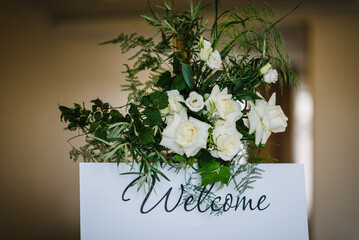 Welcome wedding on easel at reception. For seating area of guests at tables at a restaurant. The frame is decorated with white flowers and greenery. Closeup details.