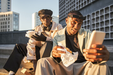 Young men in stylish clothes watching mobile phone videos and eating fast food