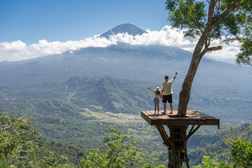 Father and daughter tourists enjoying travel around Bali island , Indonesia. They stands on photo location on the tree with view on Agung Mountain - obrazy, fototapety, plakaty