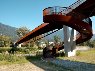 Modern rusted pedestrian and bicycle bridge with flowing Ticino river below. Detail of the spiral staircase and mountain panorama of the Swiss Alps in the background.