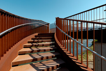 Detail of a stairway of a modern rusty bridge, with a clear blue sky behind it. Strong contrast between lights and shadows.