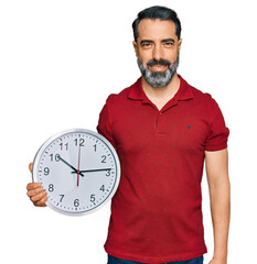 Middle aged man with beard holding big clock looking positive and happy standing and smiling with a...
