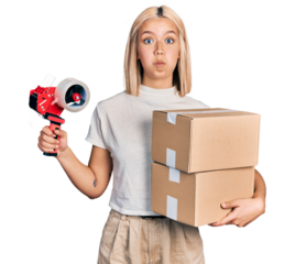 Beautiful young blonde woman holding packing tape and cardboard boxes puffing cheeks with funny face. mouth inflated with air, catching air.