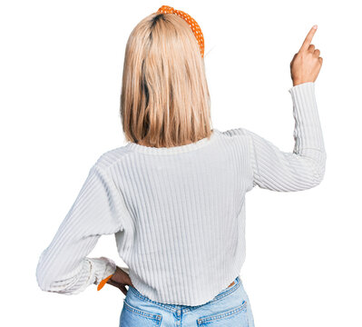 Beautiful young blonde woman wearing casual white sweater posing backwards pointing ahead with finger hand
