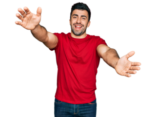 Hispanic man with beard wearing casual red t shirt looking at the camera smiling with open arms for hug. cheerful expression embracing happiness.
