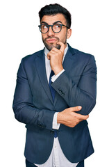 Hispanic man with beard wearing business suit and tie serious face thinking about question with hand on chin, thoughtful about confusing idea