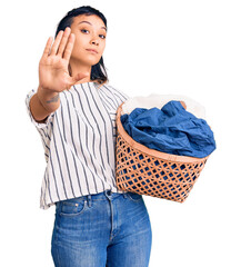 Young woman holding laundry basket with open hand doing stop sign with serious and confident...