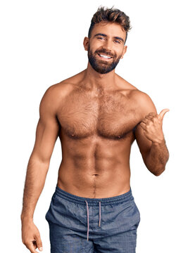 Young hispanic man wearing swimwear shirtless smiling with happy face looking and pointing to the side with thumb up.
