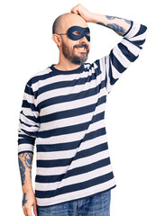 Young handsome man wearing burglar mask smiling confident touching hair with hand up gesture, posing attractive and fashionable