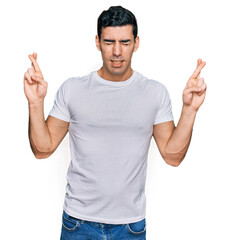 Handsome hispanic man wearing casual white t shirt gesturing finger crossed smiling with hope and eyes closed. luck and superstitious concept.