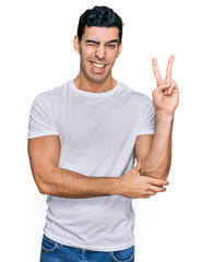 Naklejka premium Handsome hispanic man wearing casual white t shirt smiling with happy face winking at the camera doing victory sign. number two.