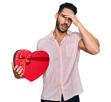 Hispanic man with beard holding valentine gift stressed and frustrated with hand on head, surprised and angry face