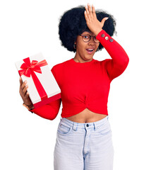 Young african american girl holding gift surprised with hand on head for mistake, remember error....