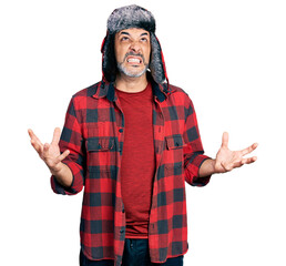Naklejka premium Middle age hispanic man with grey hair wearing fluffy earmuff hat crazy and mad shouting and yelling with aggressive expression and arms raised. frustration concept.