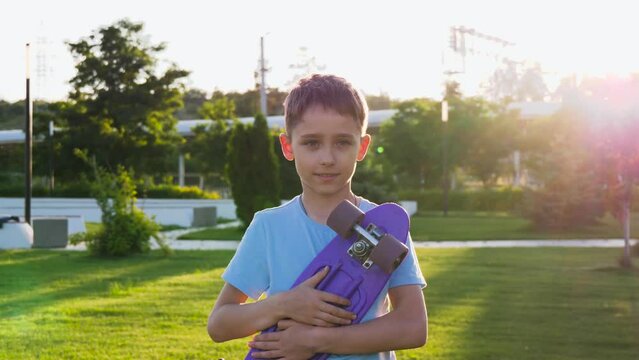 Portrait of a happy boy holding a skateboard and looking at the camera. A child with a skateboard on the background of a green park. A child rides a skateboard. Sports kid. kids dreams