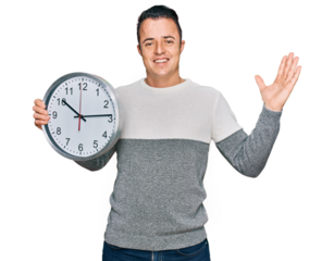 Handsome young man holding big clock celebrating victory with happy smile and winner expression with raised hands