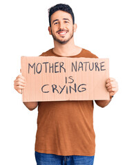 Young hispanic man holding mother nature is crying protest cardboard banner looking positive and happy standing and smiling with a confident smile showing teeth