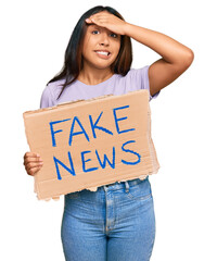 Young latin woman holding fake news banner stressed and frustrated with hand on head, surprised and...