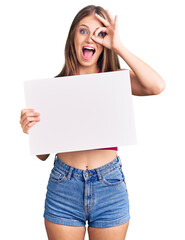 Fototapeta premium Young beautiful blonde woman holding blank empty banner smiling happy doing ok sign with hand on eye looking through fingers