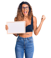 Young hispanic woman with tattoo holding empty white chalkboard screaming proud, celebrating victory and success very excited with raised arms